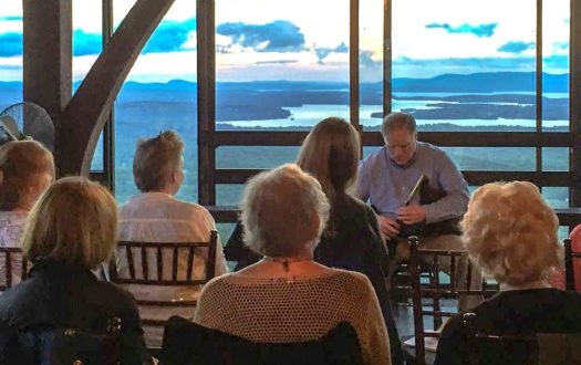 Group of people with backs to camera listen to poet in front of panoramic view of lake and mountains