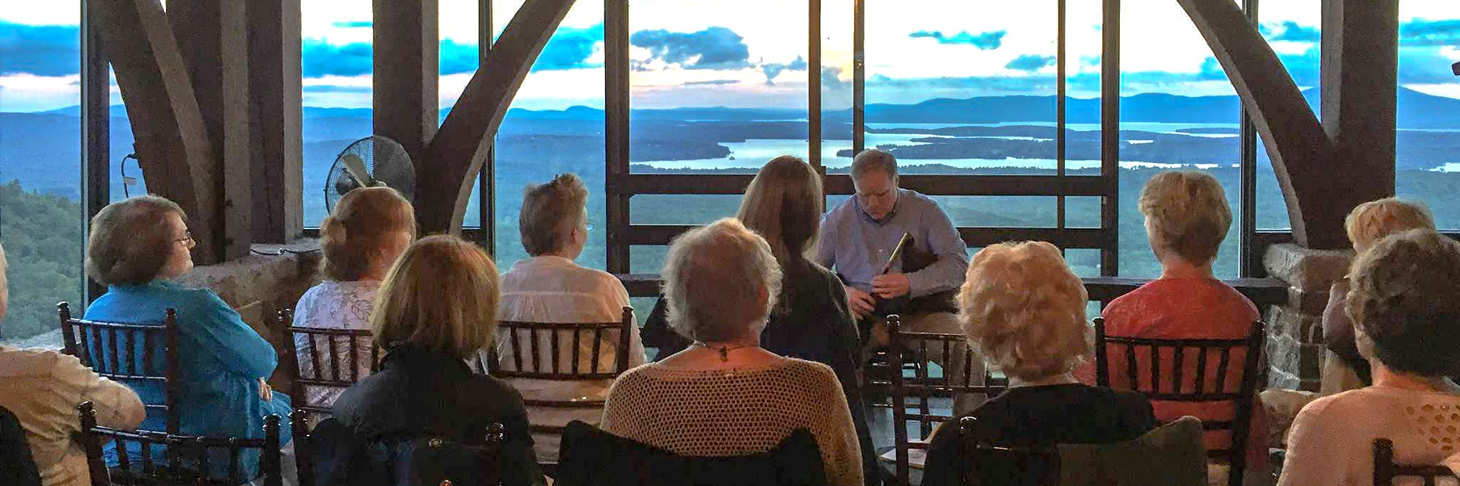 Group of people with backs to camera listen to poet in front of panoramic view of lake and mountains