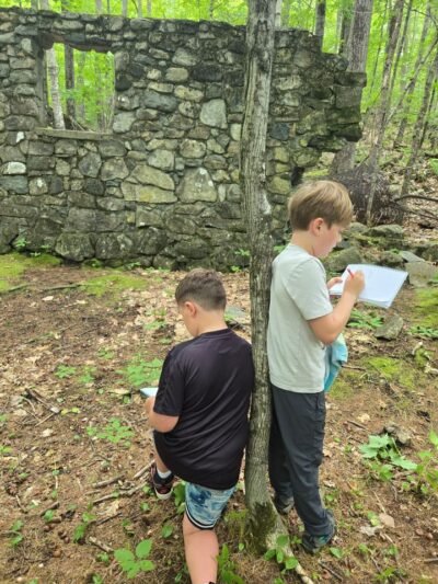 Two boys sketch in front of the ruins of a stone building as they lean back to back against a tree.